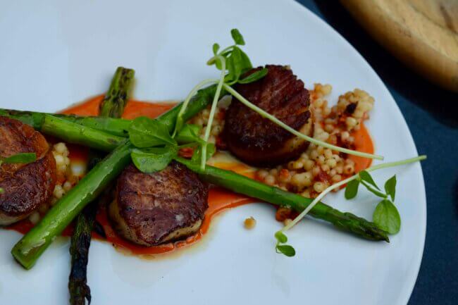 Seared scallops plated with grilled asparagus, couscous, and red sauce on a white dish.