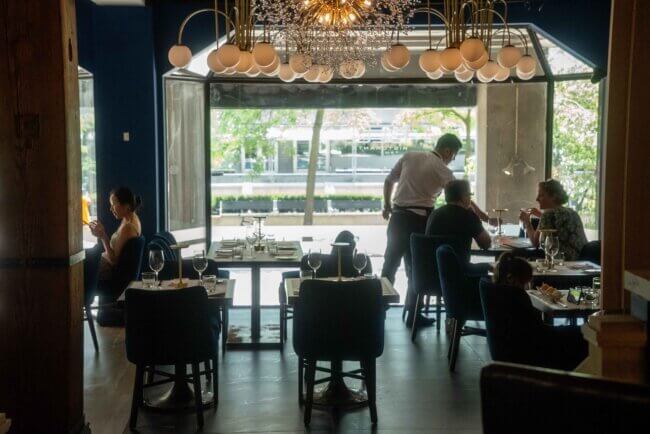 Guests dining in a bright, modern restaurant with large open windows and a server attending to a table
