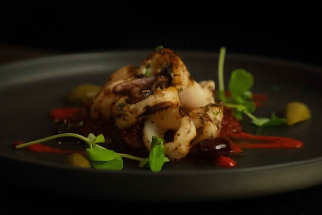 Grilled squid served on a dark plate with a drizzle of sauce and garnished with microgreens.